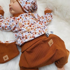 Könnte beinhalten: Eine Babypuppe trägt ein langärmeliges Oberteil und eine passende Mütze mit einem Blumenmuster in Orange, Braun und Grau auf weißem Grund. Die Puppe trägt zudem eine gerippte, rostfarbene Hose und ein passendes Lätzchen, beide mit einem kleinen Herzaufnäher.
