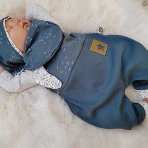 Könnte beinhalten: Ein Baby in einem blauen Outfit mit passender Kapuze und Hose. Das Outfit hat einen sternförmigen Aufnäher und ein Muster aus kleinen weißen Vögeln. Das Baby liegt auf einer flauschigen weißen Oberfläche.