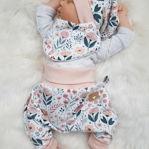 Op de afbeelding: Een baby draagt een bijpassende outfit met bloemenprint, inclusief een muts, slab en broek. De outfit heeft een witte achtergrond met roze en blauwgroene bloempatronen. De baby ligt op een witte, zachte ondergrond.