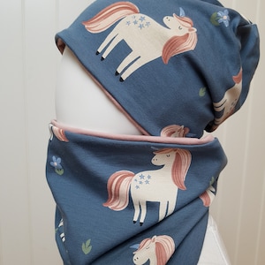 Könnte beinhalten: Eine blaue Strickmütze und ein passender Halsschmuck mit einem Einhorn-Druck. Die Einhörner sind weiß mit rosa Mähnen und Schwänzen und haben Sterne auf ihren Körpern.