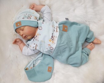 Könnte beinhalten: Ein Baby in einem hellblauen Outfit, bestehend aus Mütze, langärmligem Oberteil, Hose und Lätzchen. Das Outfit hat einen Traktor- und Tierdruck. Mütze und Hose haben einen braunen sternförmigen Aufnäher. Das Baby liegt auf einer weißen, flauschigen Oberfläche.