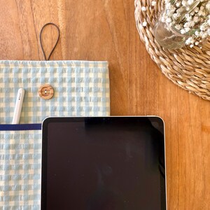 May include: A light blue and white gingham tablet sleeve with a dark blue pocket and a wooden button closure. A white stylus is in the pocket. A black tablet is in front of the sleeve. A small woven basket with white flowers is in the background.