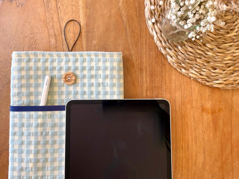 May include: A light blue and white gingham tablet sleeve with a dark blue pocket and a wooden button closure. A white stylus is in the pocket. A black tablet is in front of the sleeve. A small woven basket with white flowers is in the background.