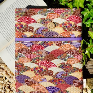 Puede incluir: Una colorida funda de libro con un diseño de abanico japonés en tonos rojos, marrones, morados y blancos. La funda tiene un cierre de botón de madera y una banda morada. El libro es parcialmente visible.