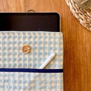 May include: A light blue and white gingham tablet sleeve with a wooden button closure. A navy blue ribbon accents the front. A white stylus pen is in the front pocket. The sleeve is on a wooden surface.
