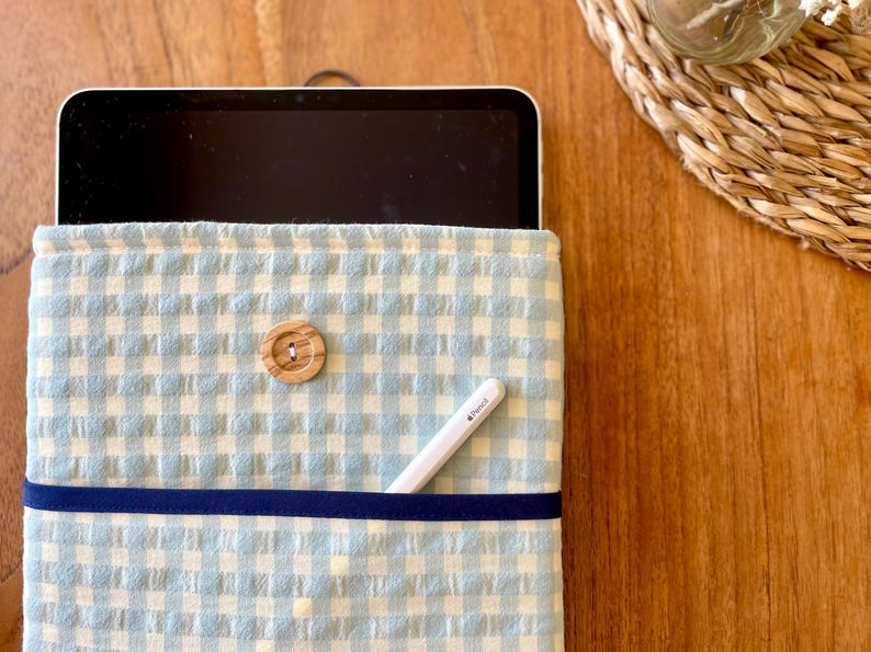 May include: A light blue and white gingham tablet sleeve with a wooden button closure. A navy blue ribbon accents the front. A white stylus pen is in the front pocket. The sleeve is on a wooden surface.