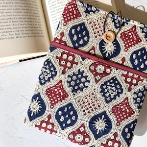 Pode incluir: Uma capa de livro estampada em tons de vermelho, azul marinho e creme, com um design repetido de formas estilizadas e elementos florais. Um botão de madeira prende a capa. A capa está aberta, revelando as páginas de um livro.