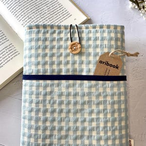 May include: A light blue and white gingham book cover with a navy blue band and a wooden button closure. A tag with the text "aribook" is attached. An open book is visible in the background.