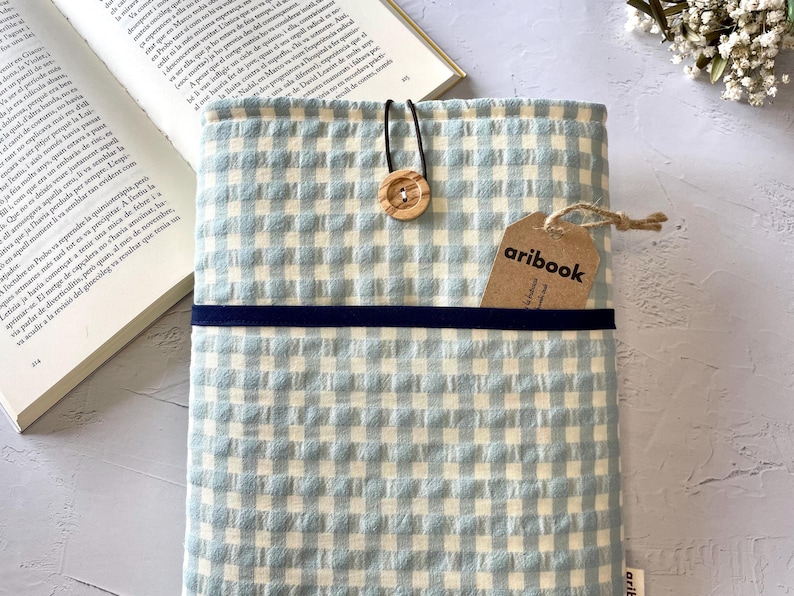 May include: A light blue and white gingham book cover with a navy blue band and a wooden button closure. A tag with the text "aribook" is attached. An open book is visible in the background.