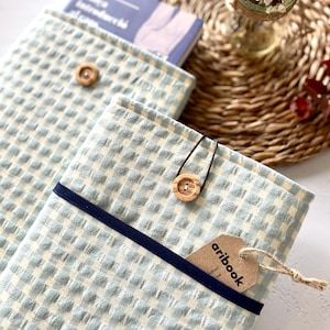 May include: Light blue and white checkered book covers with a navy blue band and wooden button closure. One cover has a tag that reads "aribook". A book is partially visible inside one cover. The covers are on a woven mat.