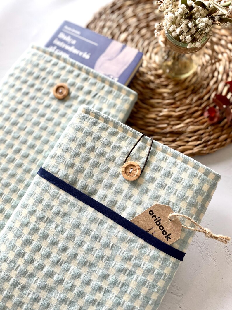 May include: Light blue and white checkered book covers with a navy blue band and wooden button closure. One cover has a tag that reads "aribook". A book is partially visible inside one cover. The covers are on a woven mat.