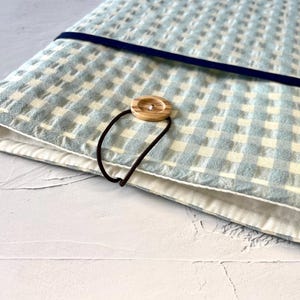 May include: A light blue and white checkered fabric case with a dark blue elastic band and a wooden button closure. The case is lined with white fabric and has a brown elastic loop.