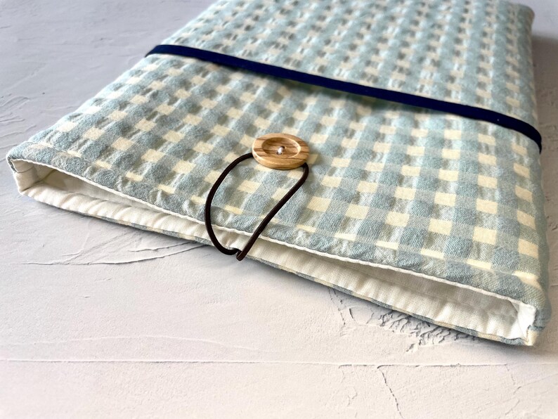 May include: A light blue and white checkered fabric case with a dark blue elastic band and a wooden button closure. The case is lined with white fabric and has a brown elastic loop.