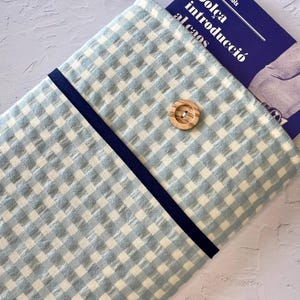May include: A light blue and white gingham fabric book sleeve with a navy blue trim and a wooden button closure. A book with text in a foreign language is partially visible inside the sleeve.