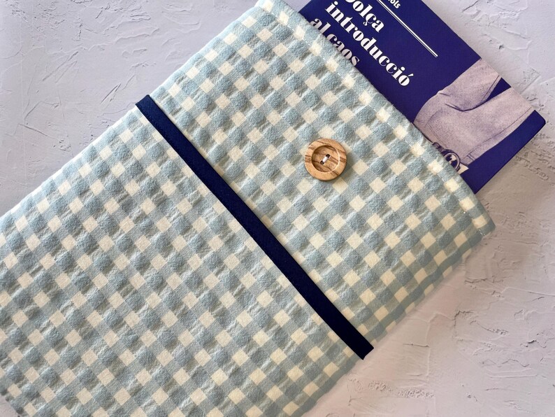 May include: A light blue and white gingham fabric book sleeve with a navy blue trim and a wooden button closure. A book with text in a foreign language is partially visible inside the sleeve.