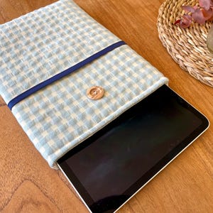 May include: A light blue and white gingham pattern tablet sleeve with a navy blue trim and a wooden button closure. The tablet is partially visible inside the sleeve. The sleeve is on a wooden surface.