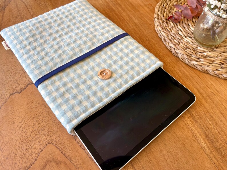May include: A light blue and white gingham pattern tablet sleeve with a navy blue trim and a wooden button closure. The tablet is partially visible inside the sleeve. The sleeve is on a wooden surface.