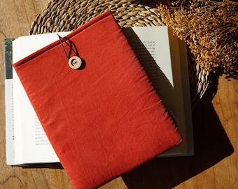 May include: A red book cover with a wooden button closure rests on an open book. The cover is made of textured fabric. The scene is set on a wooden surface with a woven mat and dried flowers.
