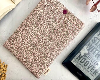 May include: A floral print Kindle cover with a button closure. The cover features a cream background with small red and gray floral patterns. A Kindle e-reader is visible next to the cover, displaying text from a book.