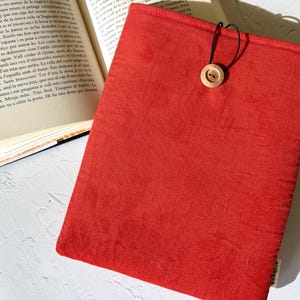 Pode incluir: Uma capa de livro de tecido vermelho com fecho de botão de madeira e cordão preto. A capa é retangular e tem uma superfície texturizada. Um livro aberto é parcialmente visível no fundo.