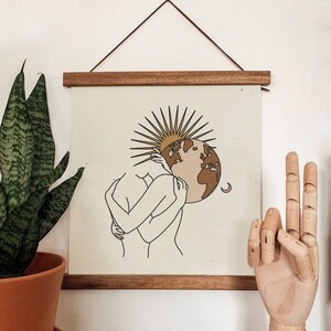 K&ouml;nnte beinhalten: Ein minimalistischer Line-Art-Druck von zwei Figuren, die sich umarmen, mit einer stilisierten Sonne und einem braunen und rosafarbenen Globus dahinter. Der Druck ist in einem hellbraunen Holzrahmen gerahmt.