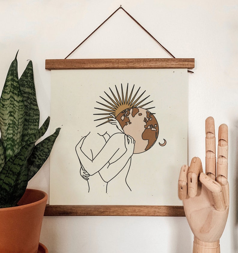 K&ouml;nnte beinhalten: Ein minimalistischer Line-Art-Druck von zwei Figuren, die sich umarmen, mit einer stilisierten Sonne und einem braunen und rosafarbenen Globus dahinter. Der Druck ist in einem hellbraunen Holzrahmen gerahmt.