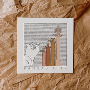 May include: A black and white illustration of a cat looking up at a row of five buildings with the text "KANSAS CITY" below. The buildings are stylized and have a retro look.