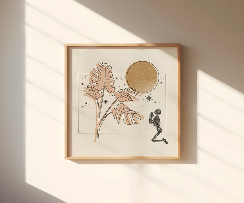 Op de afbeelding: Een ingelijste print met een minimalistisch ontwerp met een gestileerd skelet dat bidt onder een grote, gouden maan en een tropische plant met grote bladeren. De achtergrond is lichtbeige met kleine sterren.