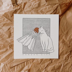 Puede incluir: Un dibujo en blanco y negro de un gato jugando con un volante de b&aacute;dminton. La imagen est&aacute; sobre un fondo a rayas y el texto "KANSAS CITY" est&aacute; impreso debajo de la imagen.