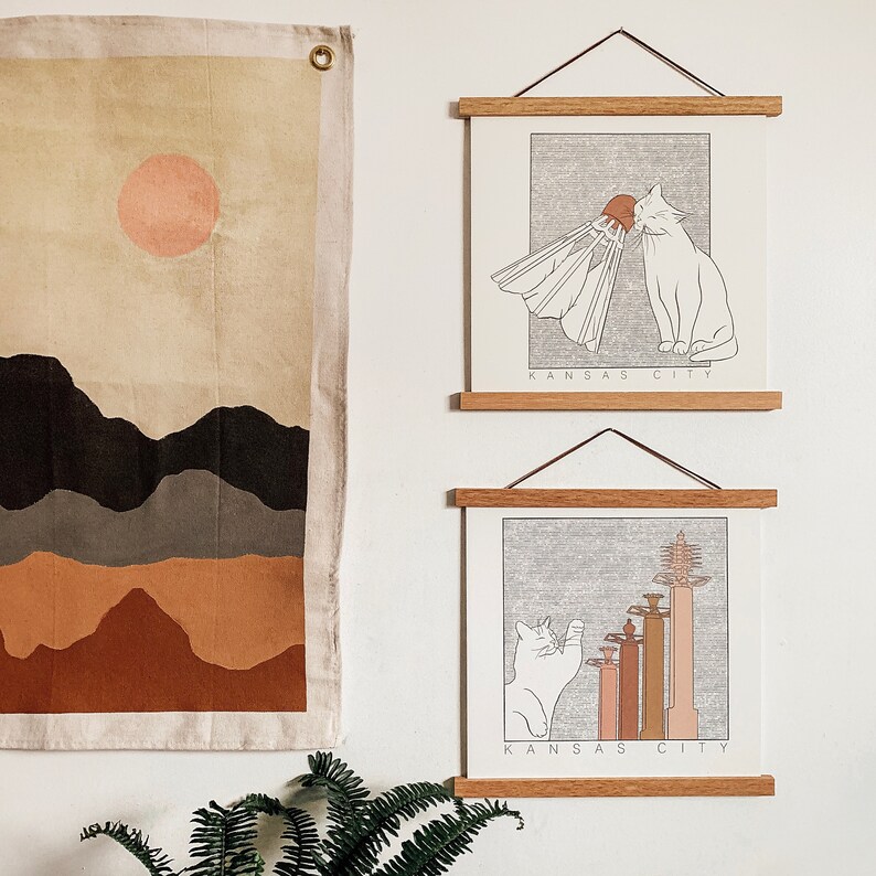 May include: Two framed prints with a white background and black line drawings of cats. The top print features a cat playing badminton with the text "KANSAS CITY". The bottom print features a cat looking at a skyline with the text "KANSAS CITY".