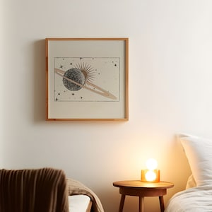 May include: A framed print of a celestial scene with a planet and rings, a sun, and stars on a beige background. The print is hanging on a white wall above a wooden nightstand with a lamp.