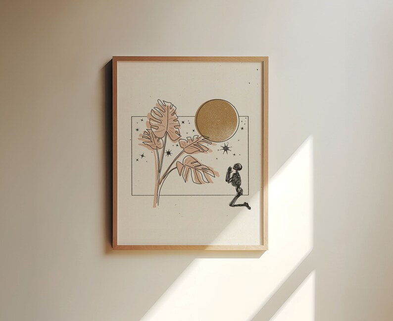 Op de afbeelding: Een ingelijst print met een minimalistische lijntekening van een plant met grote bladeren, een gouden cirkel en een knielend skeletfiguur. De achtergrond is lichtbeige met kleine zwarte sterren.