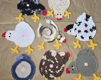 May include: A collection of handmade crochet chicken coasters in various colors, including black, white, gray, and brown. Each coaster features a circular body, red comb, yellow feet, and a pink center. The coasters are arranged on a brown surface.