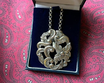May include: A silver pendant necklace with a detailed floral design. The pendant is in the shape of a circle and features intricate swirls and leaves.