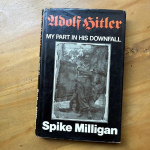 Op de afbeelding: Een hardcover boek getiteld "Adolf Hitler: My Part in His Downfall" van Spike Milligan. De omslag is zwart met witte en oranje tekst. Een zwart-wit foto van een man staat in het midden van de omslag.