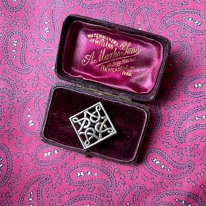 May include: A silver-colored square brooch with an intricate Celtic knot design, displayed in an open, vintage jewelry box with a burgundy velvet interior. The box is marked "Watchmakers Jewellers A. Marks & Sons, Newcastle on Tyne."