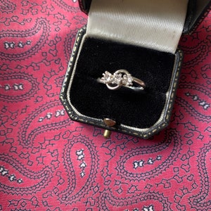 Puede incluir: Un anillo de plata con tres pequeños diamantes engastados en un diseño de nudo. El anillo está en una caja de anillo de terciopelo negro.