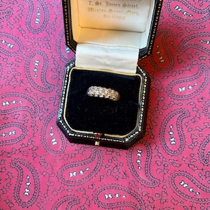 Puede incluir: Un anillo de oro con múltiples piedras pequeñas y claras, presentado en un joyero negro con ribete dorado. La caja está abierta, revelando el anillo contra un interior de terciopelo negro. El fondo es un estampado de cachemira rosa.