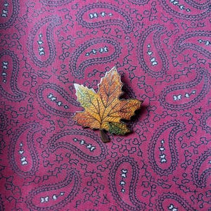 May include: A gold-toned metal brooch in the shape of a maple leaf with red, yellow, and green enamel details. The leaf is set against a pink paisley patterned fabric.