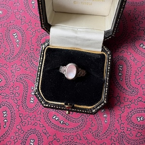 Puede incluir: Anillo de plata con una piedra preciosa ovalada de color rosa pálido, engastada en un diseño calado ornamentado. El anillo se exhibe en un joyero negro con ribete dorado, sobre un fondo rosa con estampado de cachemir.