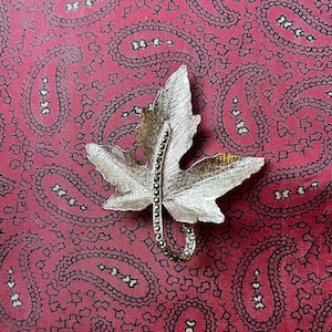 May include: A silver-toned leaf-shaped brooch with a textured surface and a curved stem adorned with small, sparkling stones. The brooch is set against a red paisley patterned background.