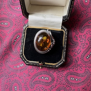 Könnte beinhalten: Ein silberner Ring mit einem ovalen Bernstein-Edelstein, präsentiert in einer schwarzen Schmuckschatulle mit weißem Satineinsatz. Der Bernstein hat eine warme, goldbraune Farbe. Die Schatulle liegt auf pinkfarbenem Paisley-Stoff.