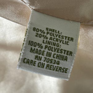 May include: A white fabric care label with green text. The label states "Shell: 80% Polyester 20% Acrylic, Lining: 100% Polyester, Made in China, RN 70534, Care on Reverse."