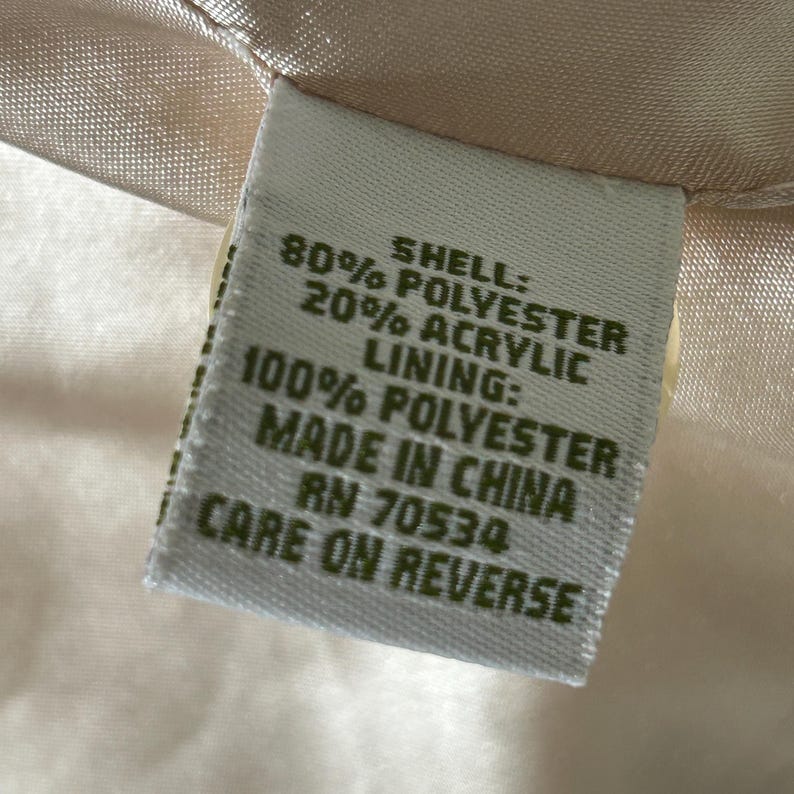 May include: A white fabric care label with green text. The label states "Shell: 80% Polyester 20% Acrylic, Lining: 100% Polyester, Made in China, RN 70534, Care on Reverse."