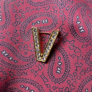 Peut inclure: Une broche en forme de lettre V dorée avec des strass transparents. La broche est sur un fond de tissu rouge à motifs.