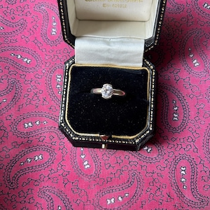 May include: A silver ring with a large, clear gemstone is presented in an open black jewellery box with a white satin interior. The box is set against a pink paisley patterned background.
