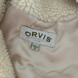 May include: A white fleece jacket with a brown Orvis label that reads "Sporting Traditions Since 1856". The label also has a smaller label that says "Made in China M".
