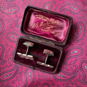 May include: A pair of silver cufflinks displayed in a vintage burgundy case with a velvet interior. The cufflinks have a rectangular design with a decorative pattern. The case is open, revealing the cufflinks and the text "A. Marks & Sons, Watchmakers, Jewellers, Newcastle on Tyne."