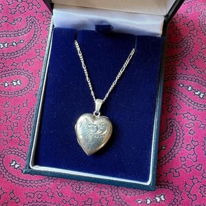 Puede incluir: Un colgante de relicario en forma de corazón de plata cuelga de una delicada cadena de plata. El relicario presenta un patrón de remolinos grabado en su superficie frontal. El collar se exhibe dentro de un joyero forrado en terciopelo azul oscuro.