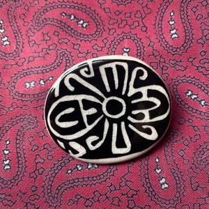 Könnte beinhalten: Eine runde Keramiknadel mit einem schwarz-weißen Design einer stilisierten Blume. Die Blume hat einen großen Mittelkreis und Blütenblätter, die aus Linien und Kurven bestehen.